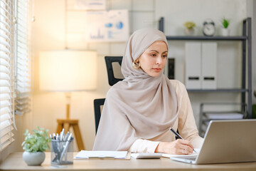A woman wearing a head scarf sits at a desk with a laptop and a pen