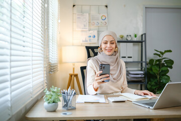 A woman wearing a hijab is sitting at a desk with a laptop and a cell phone