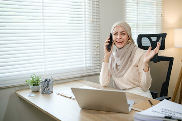 A woman wearing a hijab is talking on her cell phone while sitting at a desk