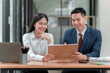 A man and a woman are sitting at a desk with a laptop and a tablet. The man is wearing a suit and tie, and the woman is wearing a white shirt. They are looking at a tablet together