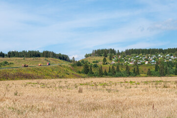 Summer rural landscape, small village on a hillside among a coniferous forest. Large yellow meadow in the foreground. A busy highway to the left of the village