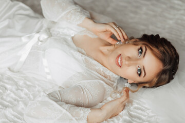 A woman is laying on a bed in a white robe. She is wearing earrings and has a smile on her face