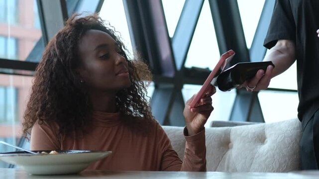 A customer uses her smartphone to make an electronic payment and pay her bill at a cafe. A very beautiful African American woman receives cashless service in a restaurant from a waiter.