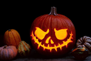 Glowing scary face carved in Halloween pumpkin among various small decorative pumpkins, jack'o lantern with burning candle inside on dark background