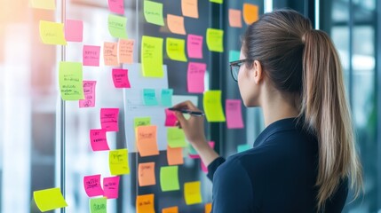Young professional woman organizes ideas with sticky notes on a glass wall, brainstorming and planning for a project in an office setting.