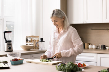 Happy mature woman spends weekend time in kitchen, cut fresh vegetables for vegetarian salad, try...