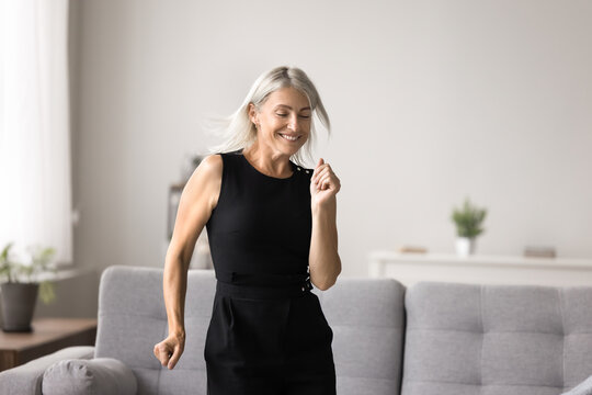 Lively middle-aged female listens favourite music and dancing in modern living room alone on weekend leisure, celebrate great news, relish life looking carefree and contented. Hobby, active lifestyle