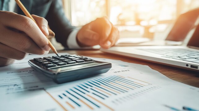 Closeup of a businessman or accountant holding a pencil while working on a calculator to calculate financial data accompanied by accountancy documents and a laptop in an office setting