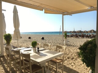 The image shows a beachfront dining area with a table and chairs under a canopy. Umbrellas and lounge chairs are set up on the sandy beach, with a view of the calm blue sea in the background.