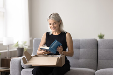 Smiling mature woman sitting on sofa unpacking box, get package, opening received parcel with...