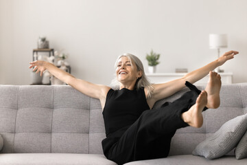 Cherry mature woman jumps on sofa with raised arms and legs enjoy comfort and holidays in modern...