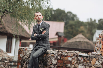 A man in a suit is sitting on a stone wall. He is wearing a tie and a flower in his lapel. The man is looking off into the distance