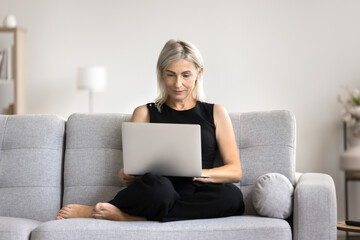 Mature woman working on laptop at home, sit on sofa with wireless computer, engaged in telework activity, buying online goods, making order using electronic commerce e-services or marketplace websites