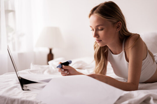 Blonde girl in white t-shirt top working with documents using laptop on bed in bedroom at home