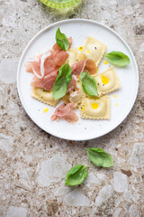 White plate with ravioli, prosciutto and fresh basil, above view on a light-brown granite background, vertical shot