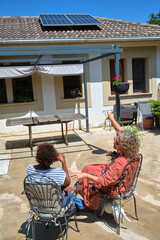Two women sitting in a sunny backyard, pointing at solar panels