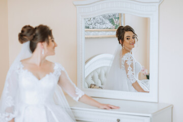 A bride is standing in front of a mirror, wearing a white dress and a veil. She is smiling and looking at her reflection. Concept of happiness and excitement, as the bride prepares for her wedding day