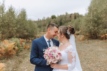 A bride and groom are standing in a field, smiling at the camera. The bride is holding a bouquet of...