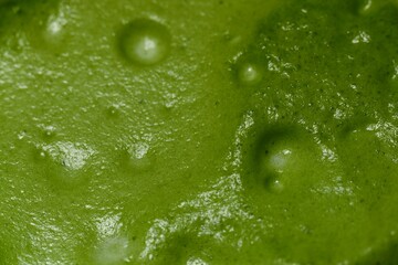 A close-up image of green tea with a smooth, frothy texture. The vibrant green color and visible air bubbles on the surface highlight the freshness and quality of the matcha