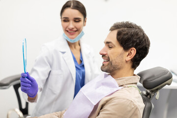 Professional female orthodontist holding mirror and showing result of teeth treatment to man in dentist clinic, side view