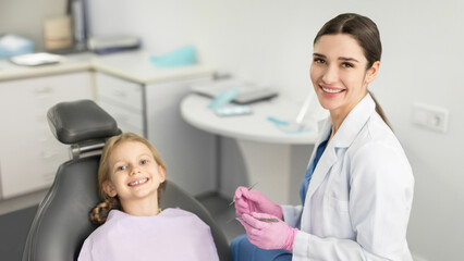 Fototapeta premium Happy child girl sitting in dental chair beside his female dentist and smiling together at camera, panorama shot