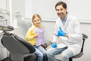 Fototapeta premium Dentistry and childcare concept. Happy man dentist and kid girl patient showing thumbs up and smiling at camera at dental clinic