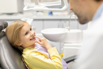 Fototapeta premium Adorable little girl showing her baby teeth to male pediatrics dentist and pointing to her smile, sitting in chair in clinic office