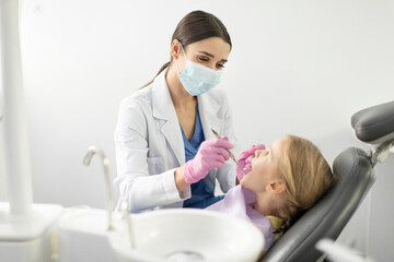Obraz premium Professional female dentist with dental instruments examining teeth of little girl patient in dental clinic