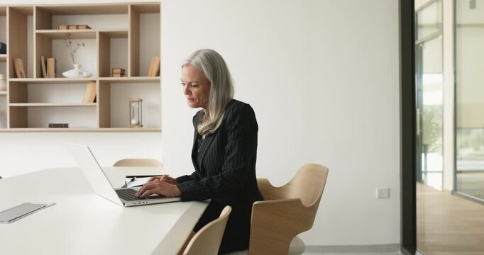 Focused senior businesswoman typing on laptop at large white table, working on online project using professional application for job communication, speaking at computer. Side view