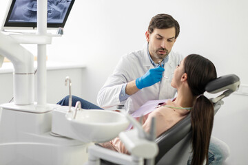 Professional male dentist examining young female patient's teeth in modern dentistry office, free space. Teeth checkup concept