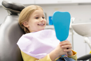 Happy lovely little European girl looking in mirror at her teeth after teeth polishing procedure in...