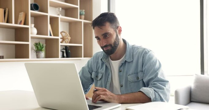 Positive Hispanic businessman in casual working at laptop from home office, writing notes, sitting at workplace table. Adult student watching webinar, using online learning app on computer