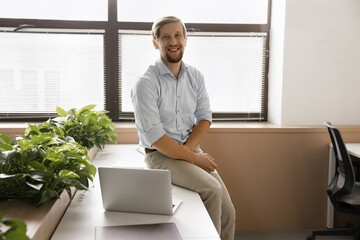 Young handsome male office employee sits on table in modern co-working space with laptop, smile, look at camera. Portrait of department chief, financial analyst or consultant, trader profile picture