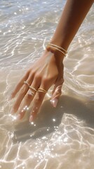 A close-up of a hand adorned with jewelry gently touching shimmering water at the beach