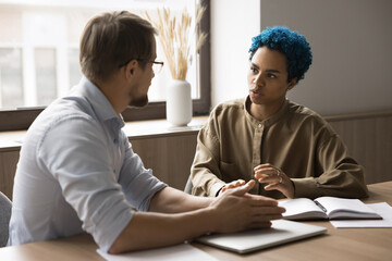 Attractive blue-haired African employee lead talk to male colleague seated at desk in office, looks...