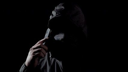 Phone scammers: a figure in a black hoodie holding a phone in the shadows. Covert cyber theft and fraud, with frightening and unseen attacks on privacy.