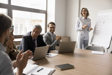 Obraz premium Laughing businesspeople gathered in boardroom, take part in educational seminar, meeting briefing event led my happy mature businesslady, company boss or team leader making flip chart presentation