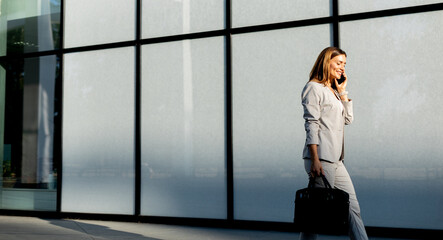 Business woman strolls confidently outdoors in smart attire, checking her messages against the backdrop of a modern glass building on a sunny day