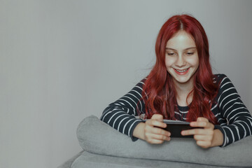 Fototapeta premium Cheerful happy girl with red hair playing games on her phone and smiling, browsing internet or communicating, emotional lifestyle portrait with copy space, idea of comfortable leisure, rest and relax