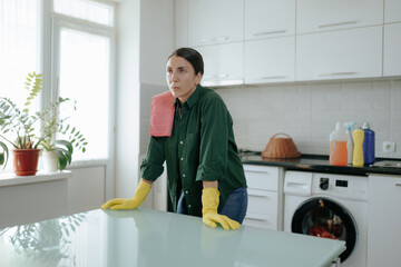 A tired young housewife with yellow gloves rests in her kitchen, taking a break from housework