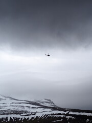 A helicopter flies over a rugged snowy mountain landscape under a dark and brooding sky, highlighting a sense of adventure and exploration in a harsh environment.