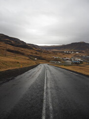 A wet road leads to a small town nestled among hills and mountains, under a gray sky, with power lines and utility poles seen along the route, capturing a rustic scene.