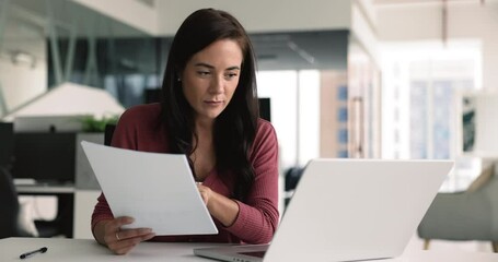 Pretty secretary holds sheets, using laptop, preparing presentation for seminar, review spreadsheets, entering information, typing, working on financial documents such as invoices, receipt, statement