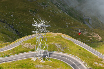 A red cable car ascends the Fagaras mountains to Lake Balea while passing by the scenic Transfăgărășan road (DN7C) that winds through the landscape underneath it, Transylvania, Romania
