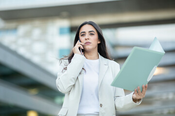 Fototapeta premium A woman in a light suit engages in a phone conversation while holding a file, standing outside a modern building.