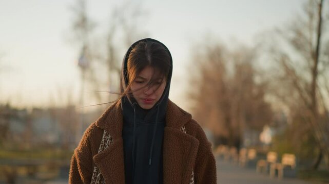Front shot of a sad girl walking through a lonely park at sunset, wearing a brown coat and a hoodie. melancholic mood with the girl's downcast expression and the serene, empty park setting