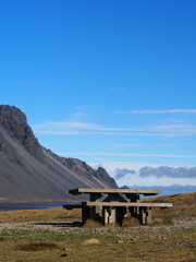 Obraz premium A wooden picnic bench set against a backdrop of rugged mountains and a calm ocean under a bright blue sky, signifying serenity and solitude.