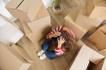 Young woman overwhelmed in her new house surrounded by cardboard boxes that reach the ceiling. Exhausted and stressed young girl opening moving boxes in student rental apartment.