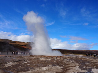 A geyser in Iceland erupts, propelling a powerful column of steam and water into the bright blue sky, surrounded by scenic and awe-inspiring landscape.
