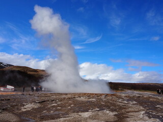Tourists witness the awe-inspiring eruption of a geyser against the backdrop of a clear blue sky, showcasing nature's power and beauty in a geothermal hotspot.